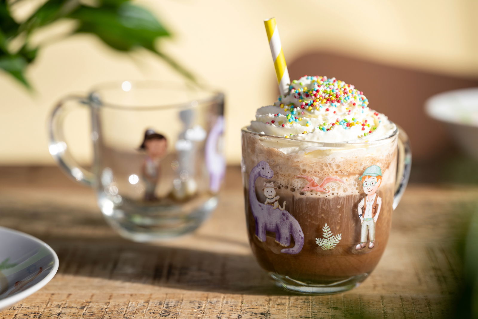 Kindertasse mit buntem Dinosaurier- und Kinderdesign, gefüllt mit Schokoladengetränk, Sahne und Streuseln, seitliche Perspektive