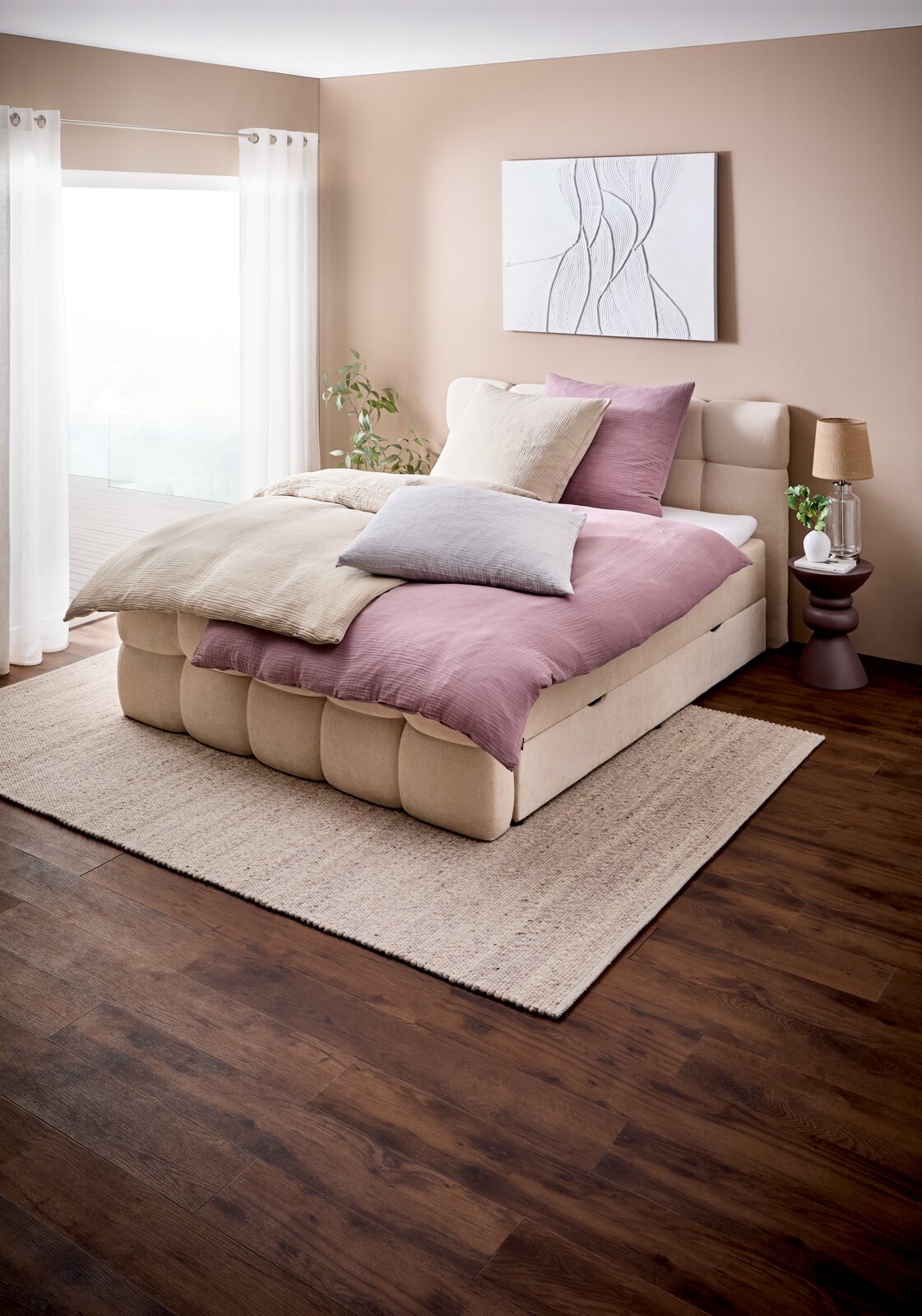 Beiges, gepolstertes Boxspringbett mit rosa und beiger Bettwäsche und mehreren Kissen im modernen Schlafzimmer, seitliche Frontansicht aus leicht erhöhter Perspektive.
