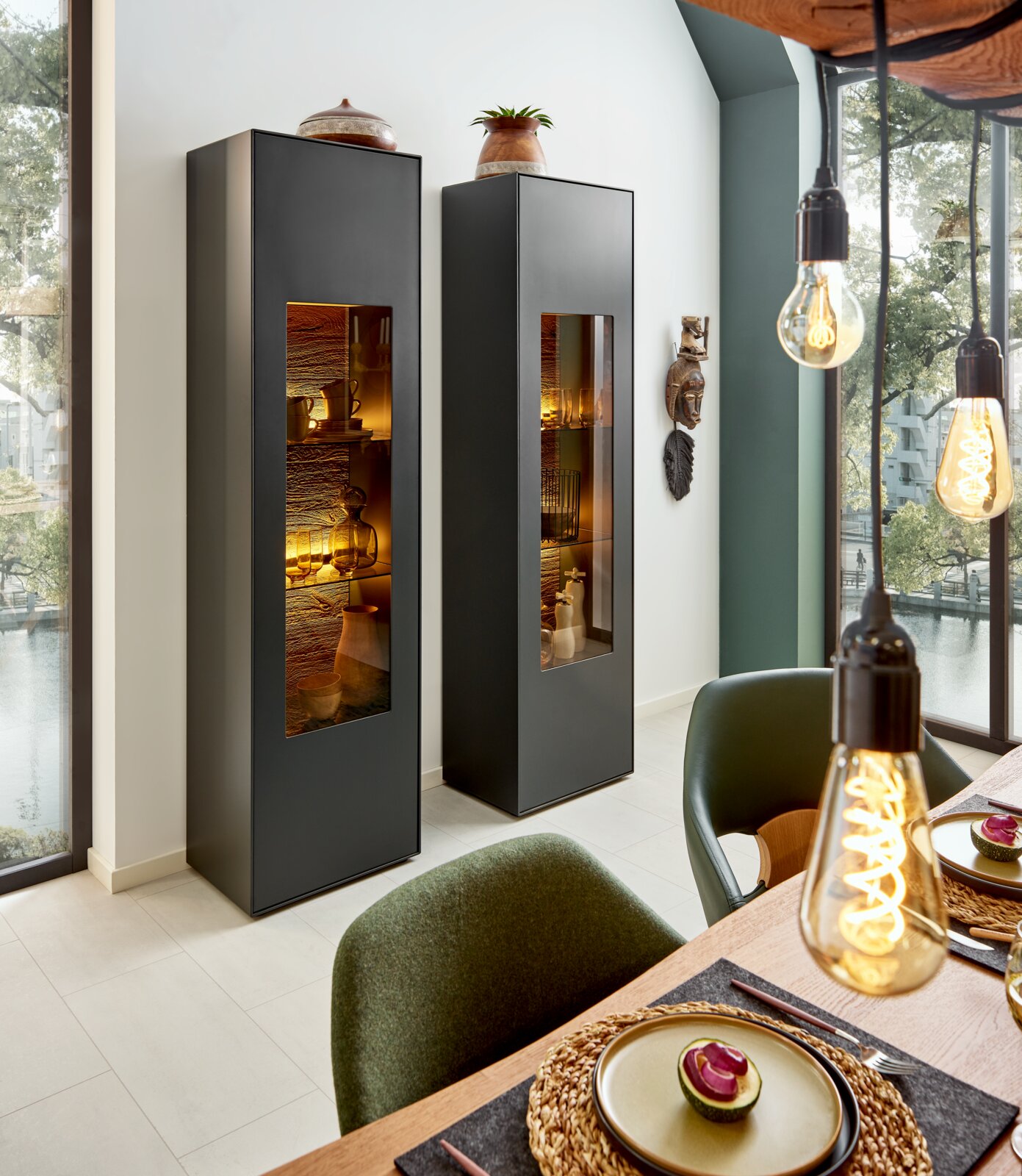 Twee moderne, zwarte vitrinekasten met glazen deuren, die vanaf de zijkant te zien zijn in een stijlvolle eetkamer met decoratieve elementen en warme verlichting.