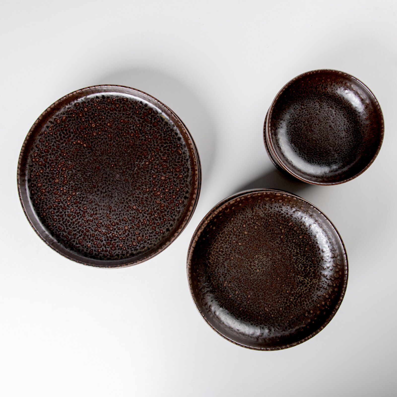 Dreiteiliges Keramikgeschirr-Set in dunklem Braun mit gesprenkelter Glasur (großer Teller, kleiner Teller und Schale), fotografiert in Draufsicht von oben auf weißem Hintergrund
