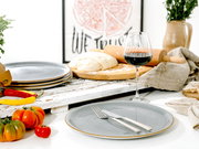 Gedeckter Tisch mit grauen Keramiktellern, Besteck und Rotweinglas neben Teig auf Holzbrett, Nudelholz und frischen Tomaten, aus leicht seitlicher Frontperspektive