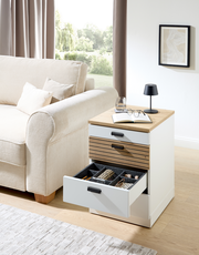 Rollcontainer GLENN mit offener Schublade, seitliche Perspektive, neben einem Sofa platziert, mit Dekorationen wie Buch, Brille und Lampe auf der Oberseite.