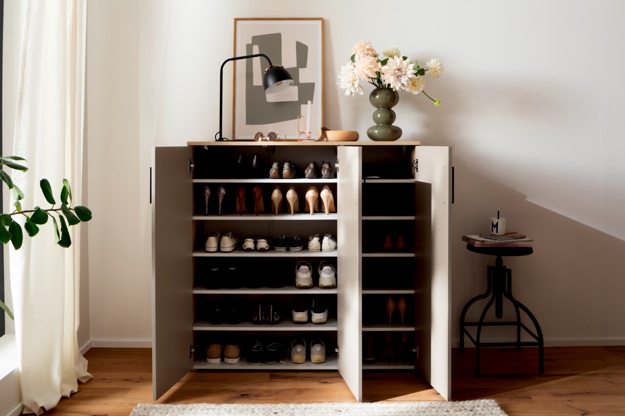 Schuhschrank breit mit offenen Türen, gefüllt mit verschiedenen Schuhen, von vorne betrachtet. Dekoriert mit einer Lampe, einer Vase mit Blumen und einem Bild auf der Oberseite.