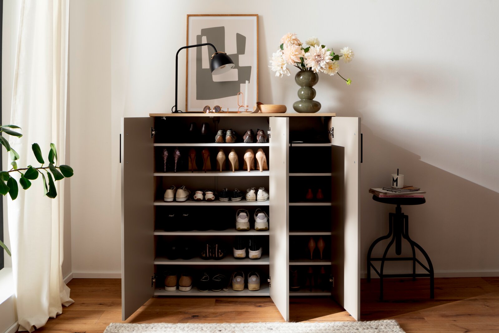 Schuhschrank breit mit offenen Türen, gefüllt mit verschiedenen Schuhen, von vorne betrachtet. Dekoriert mit einer Lampe, einer Vase mit Blumen und einem Bild auf der Oberseite.
