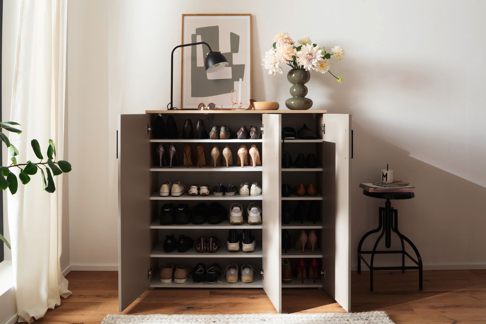 Schuhschrank breit mit offenen Türen, gefüllt mit verschiedenen Schuhen, von vorne betrachtet. Dekoriert mit einer Lampe, einer Vase mit Blumen und einem Bild auf der Oberseite.