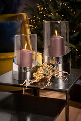 Zwei elegante Windlichter aus Glas und Metall mit brennenden Kerzen, dekoriert mit goldenen Beerenzweigen, auf einem Tisch vor einem festlich geschmückten Hintergrund. Perspektive: seitlich.