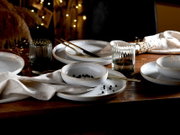 Rustikales Tischgedeck mit weißen Keramiktellern und -schalen, goldfarbenem Besteck, grauen Trinkgläsern und Leinenservietten auf Holztisch, aus leicht seitlicher Perspektive aufgenommen