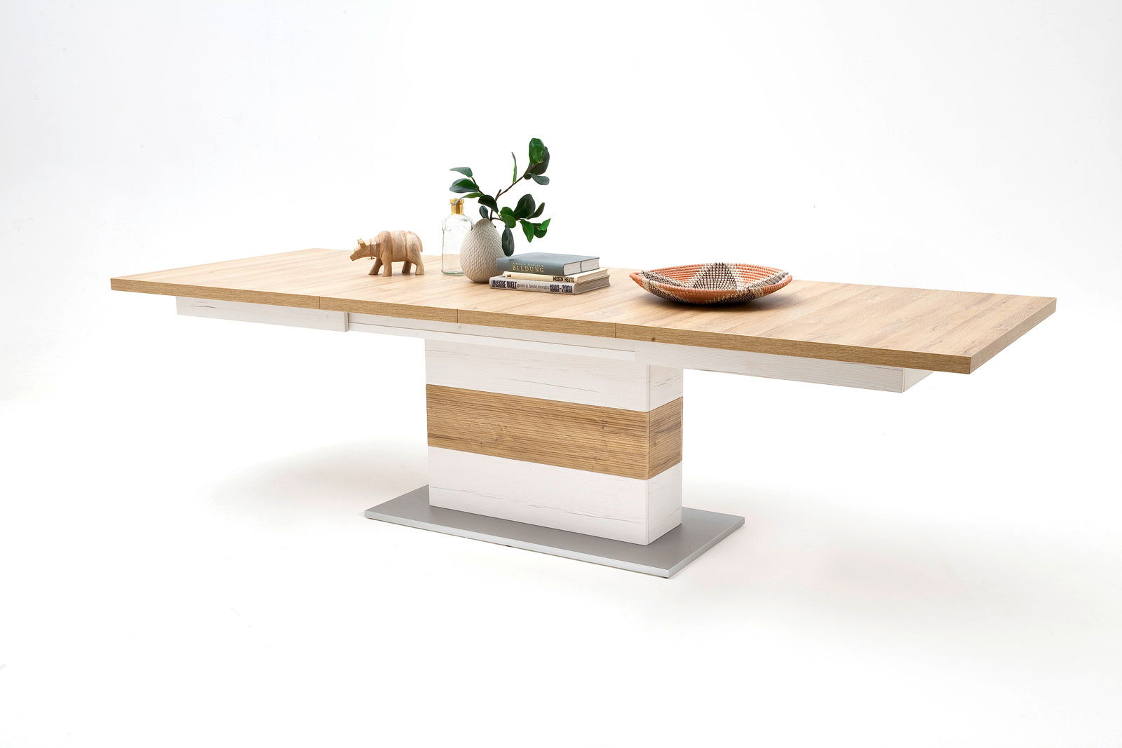 Moderner Esstisch aus Holz mit zentralem Standfuß, dekoriert mit Büchern, einer Vase und einem kleinen Holzfigur, seitliche Perspektive.