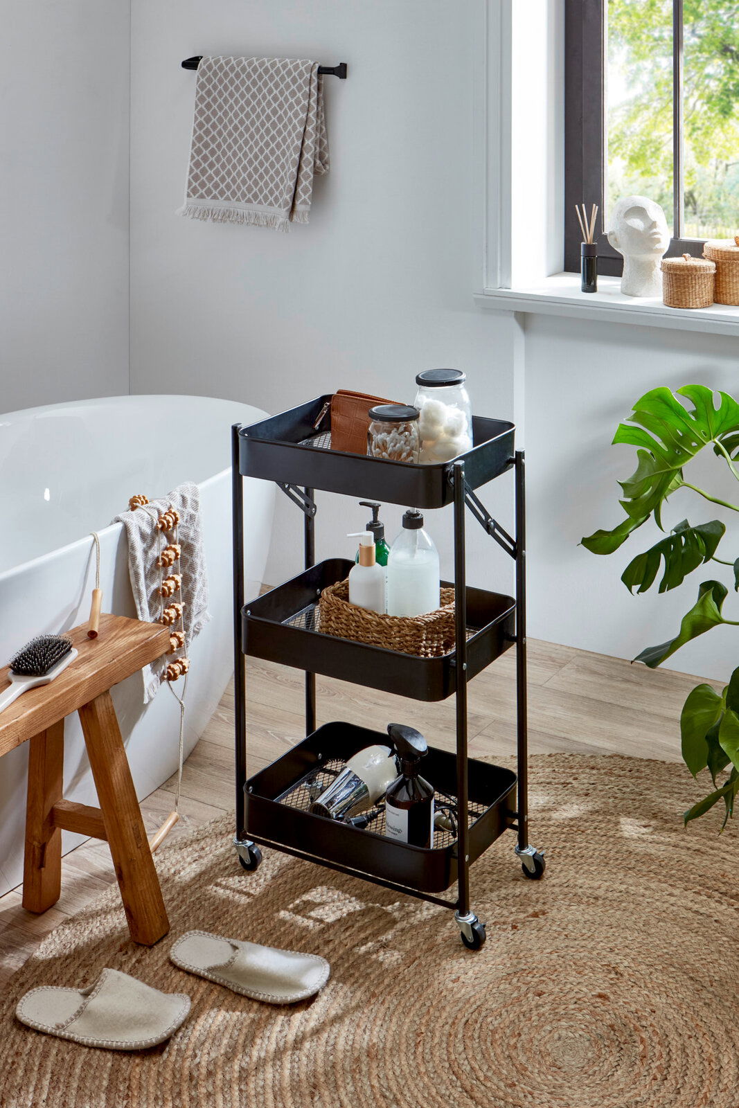 Schwarzer, dreistöckiger Metall-Rollwagen mit Ablagekörben im Badezimmer neben Badewanne und Fenster, gefüllt mit Pflege- und Badutensilien, Frontansicht in leichter Schrägperspektive von vorn.