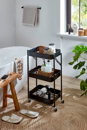 Schwarzer, dreistöckiger Metall-Rollwagen mit Ablagekörben im Badezimmer neben Badewanne und Fenster, gefüllt mit Pflege- und Badutensilien, Frontansicht in leichter Schrägperspektive von vorn.