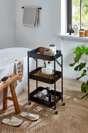 Schwarzer, dreistöckiger Metall-Rollwagen mit Ablagekörben im Badezimmer neben Badewanne und Fenster, gefüllt mit Pflege- und Badutensilien, Frontansicht in leichter Schrägperspektive von vorn.