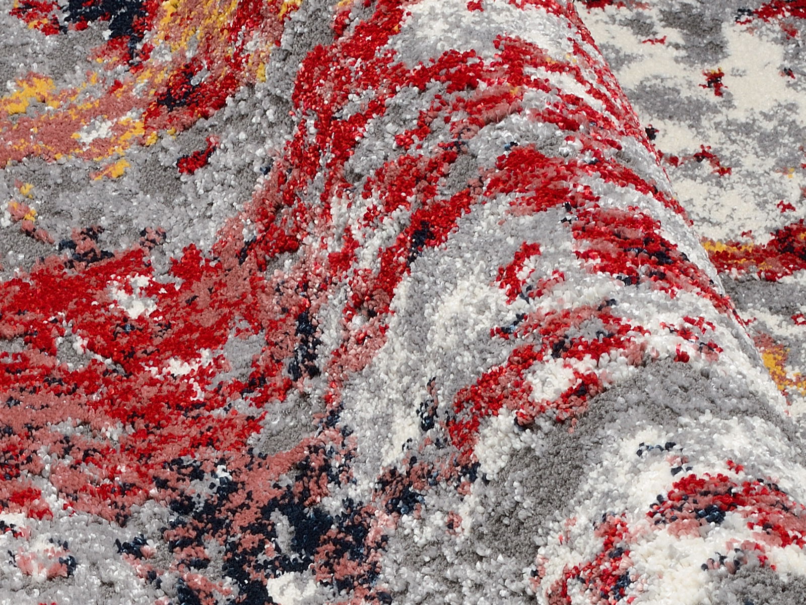 Nahaufnahme eines Designer-Teppichs mit abstraktem Muster in Rot-, Grau- und Gelbtönen, aus seitlicher Perspektive aufgenommen.