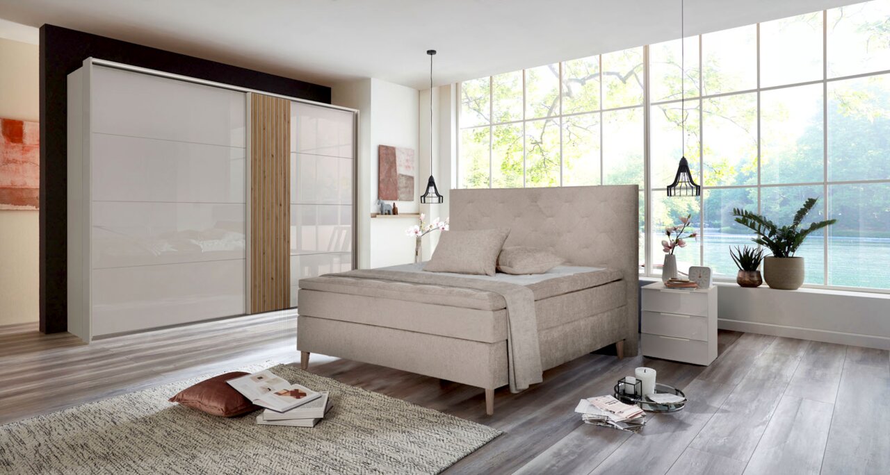 Modernes Schlafzimmer mit einem beigen Boxspringbett und einem zweiflügeligen Schwebetürenschrank, seitliche Perspektive.