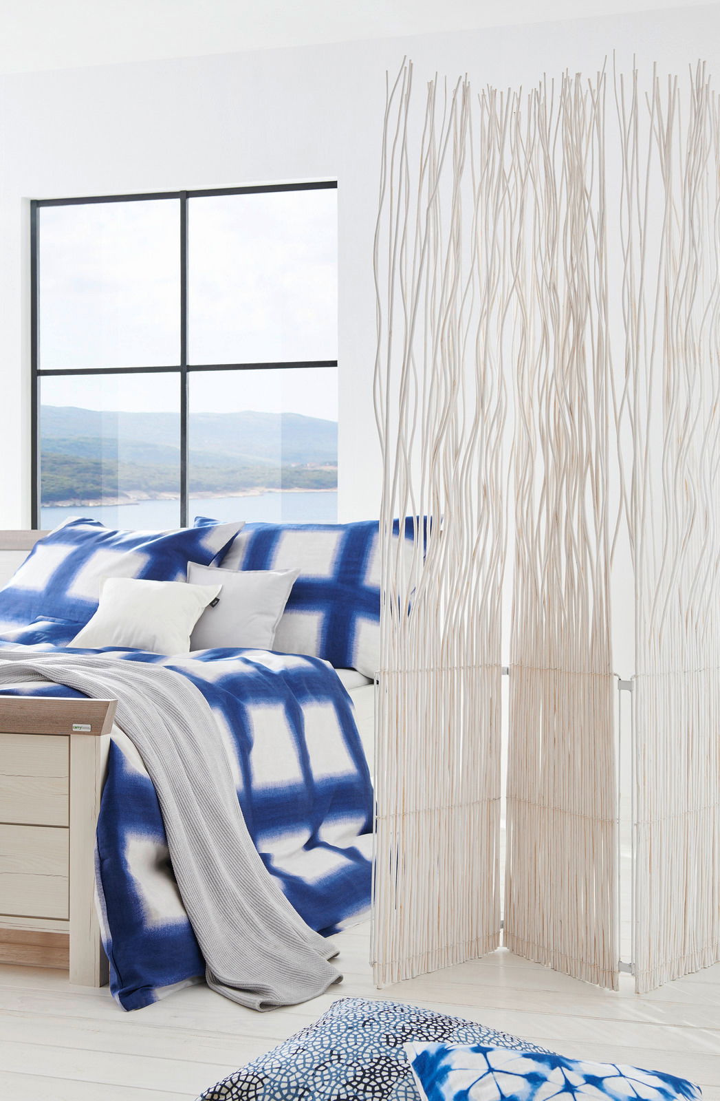Modernes Schlafzimmer mit einem Bett 180x200, das mit blauer und weißer Bettwäsche dekoriert ist. Im Vordergrund steht ein dekorativer Raumteiler aus weißen Zweigen. Die Perspektive zeigt das Bett von der Seite mit einem Blick auf ein großes Fenster im Hi