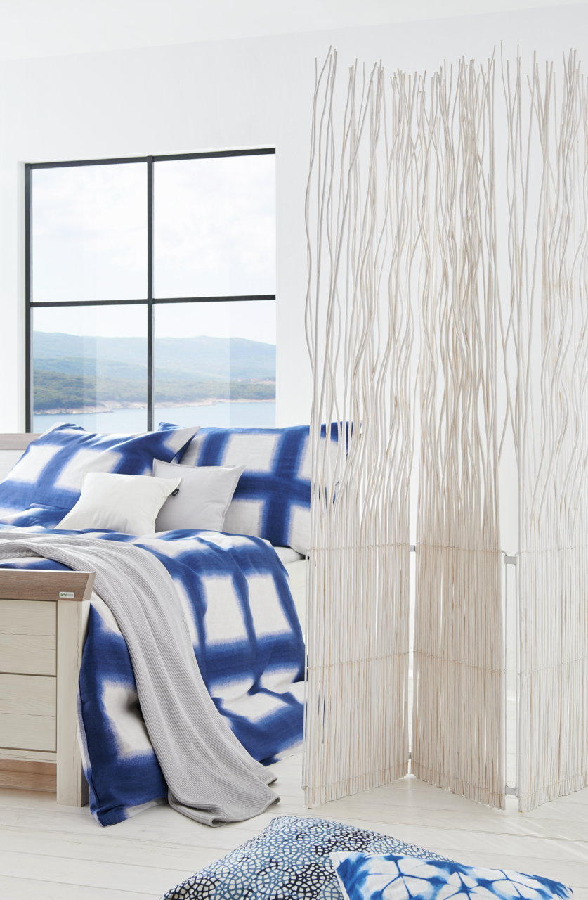 Modernes Schlafzimmer mit einem Bett 180x200, das mit blauer und weißer Bettwäsche dekoriert ist. Im Vordergrund steht ein dekorativer Raumteiler aus weißen Zweigen. Die Perspektive zeigt das Bett von der Seite mit einem Blick auf ein großes Fenster im Hi