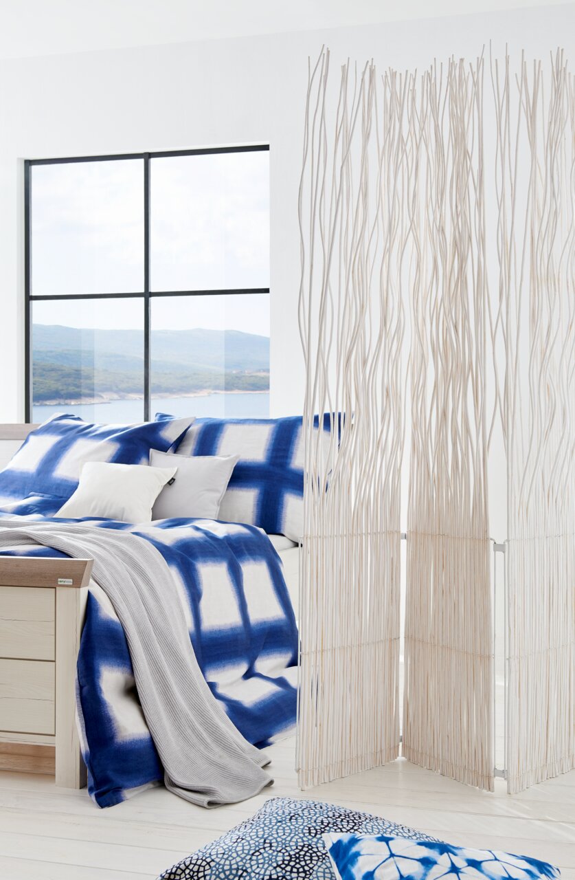 Modernes Schlafzimmer mit einem Bett 180x200, das mit blauer und weißer Bettwäsche dekoriert ist. Im Vordergrund steht ein dekorativer Raumteiler aus weißen Zweigen. Die Perspektive zeigt das Bett von der Seite mit einem Blick auf ein großes Fenster im Hi