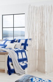 Modernes Schlafzimmer mit einem Bett 180x200, das mit blauer und weißer Bettwäsche dekoriert ist. Im Vordergrund steht ein dekorativer Raumteiler aus weißen Zweigen. Die Perspektive zeigt das Bett von der Seite mit einem Blick auf ein großes Fenster im Hi