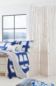 Modernes Schlafzimmer mit einem Bett 180x200, das mit blauer und weißer Bettwäsche dekoriert ist. Im Vordergrund steht ein dekorativer Raumteiler aus weißen Zweigen. Die Perspektive zeigt das Bett von der Seite mit einem Blick auf ein großes Fenster im Hi