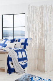 Modernes Schlafzimmer mit einem Bett 180x200, das mit blauer und weißer Bettwäsche dekoriert ist. Im Vordergrund steht ein dekorativer Raumteiler aus weißen Zweigen. Die Perspektive zeigt das Bett von der Seite mit einem Blick auf ein großes Fenster im Hi
