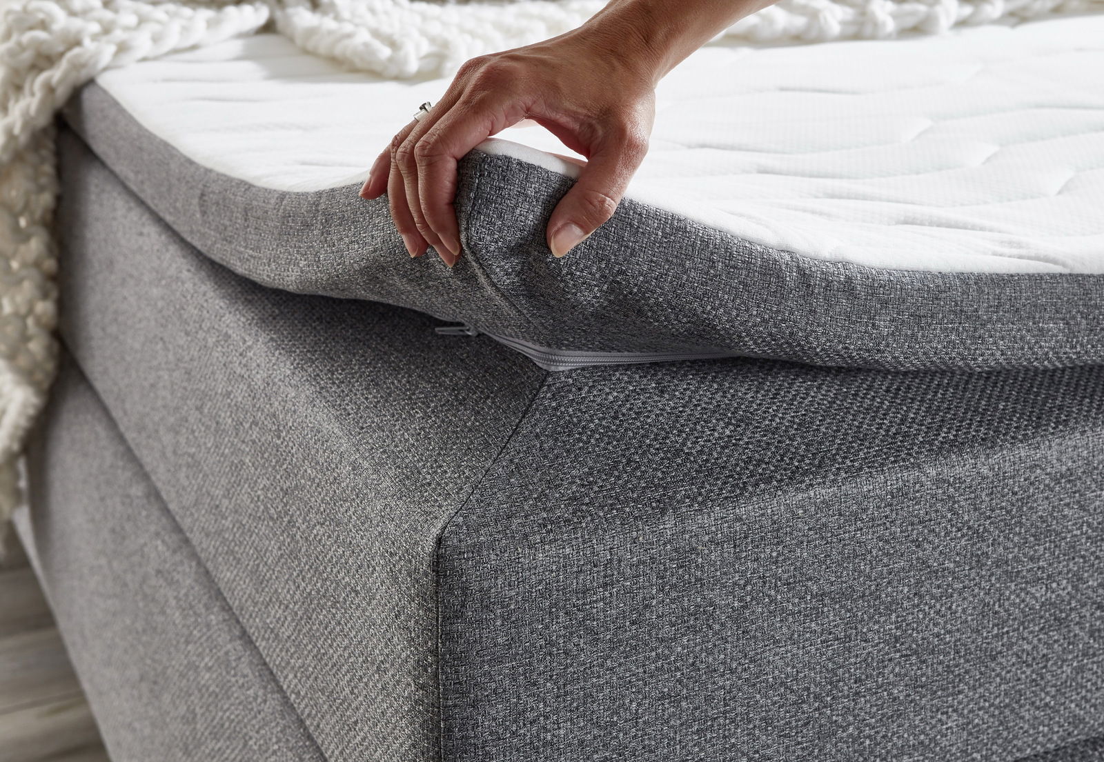 Nahaufnahme eines grauen Boxspringbetts mit einer Hand, die die Matratze anhebt, um die Struktur und den Reißverschluss zu zeigen.