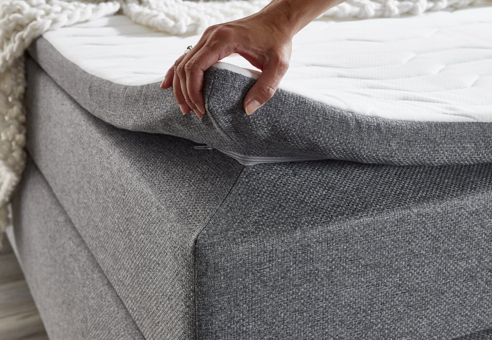 Nahaufnahme eines grauen Boxspringbetts mit einer Hand, die die Matratze anhebt, um die Struktur und den Reißverschluss zu zeigen.