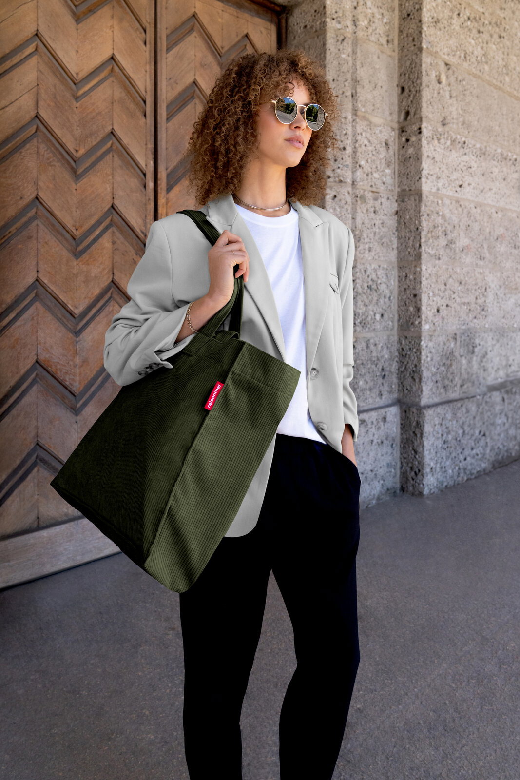 Frau mit Sonnenbrille trägt eine olivgrüne Cord-Shopper-Tasche mit rotem Label über der Schulter, aufgenommen in leichter Untersicht aus schräger Frontperspektive vor einer Holztür und Steinwand.