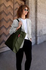Frau mit Sonnenbrille trägt eine olivgrüne Cord-Shopper-Tasche mit rotem Label über der Schulter, aufgenommen in leichter Untersicht aus schräger Frontperspektive vor einer Holztür und Steinwand.