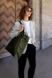 Frau mit Sonnenbrille trägt eine olivgrüne Cord-Shopper-Tasche mit rotem Label über der Schulter, aufgenommen in leichter Untersicht aus schräger Frontperspektive vor einer Holztür und Steinwand.