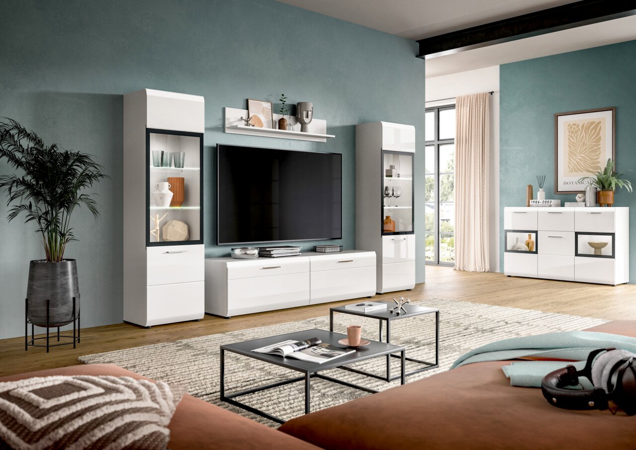 Modernes weißes Wohnzimmermöbel-Set mit TV-Lowboard, zwei Vitrinen und Sideboard vor petrolfarbener Wand, frontal aus leicht schräger Perspektive aufgenommen.