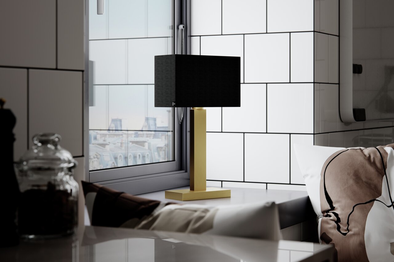 Moderne tafellamp PRIME met zwarte kap en goudkleurige voet, zijdelings op een tafel voor een raam geplaatst.
