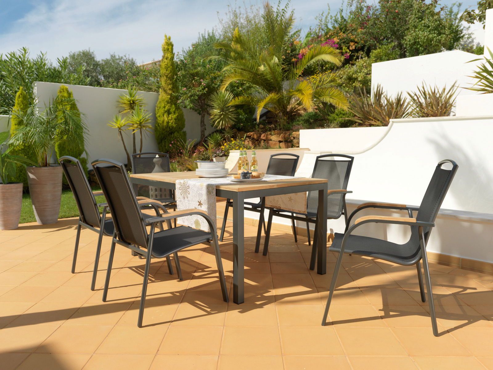 Ambia Garden Stapelsessel PLANO Moderner Gartentisch mit sechs schwarzen Stühlen auf einer sonnigen Terrasse, umgeben von grünen Pflanzen, aus seitlicher Perspektive.