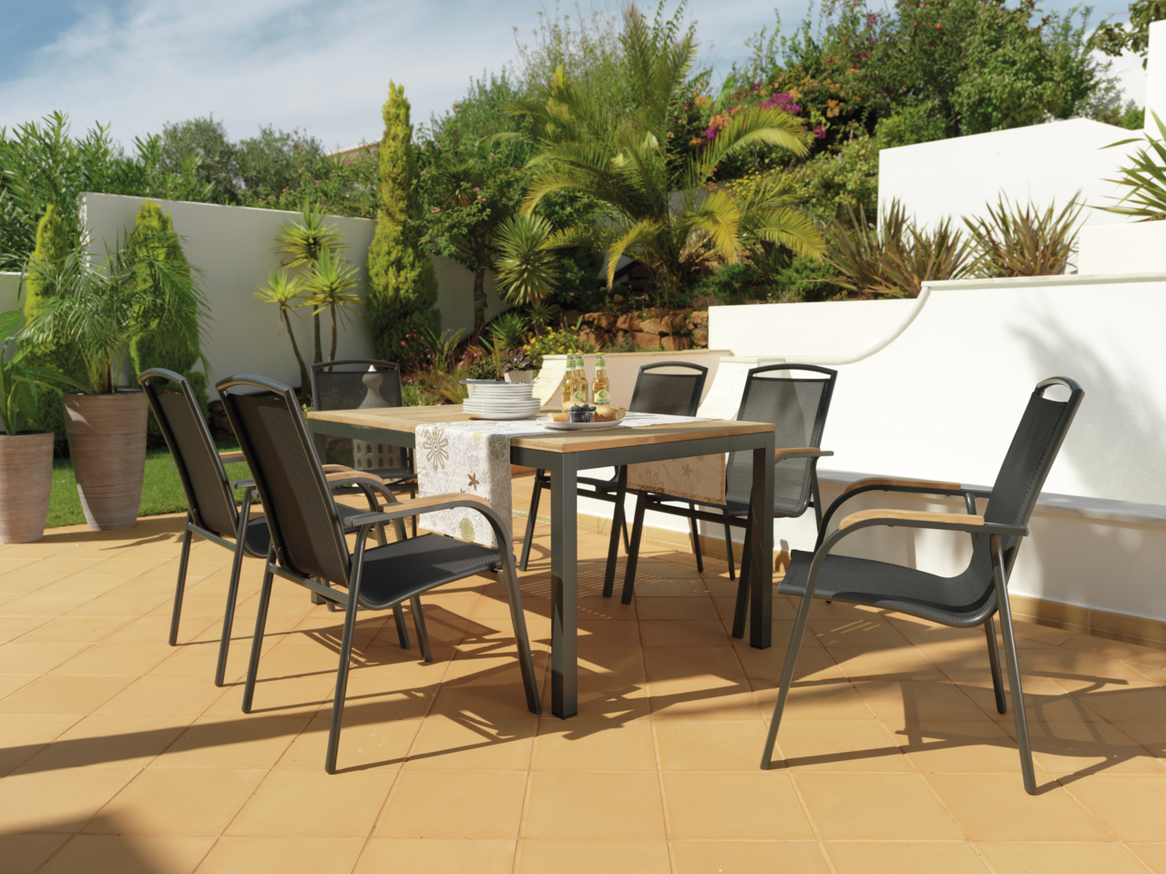 Ambia Garden Ausziehtisch PLANO Moderner Gartentisch mit sechs schwarzen Stühlen auf einer sonnigen Terrasse, umgeben von grünen Pflanzen, aus seitlicher Perspektive.