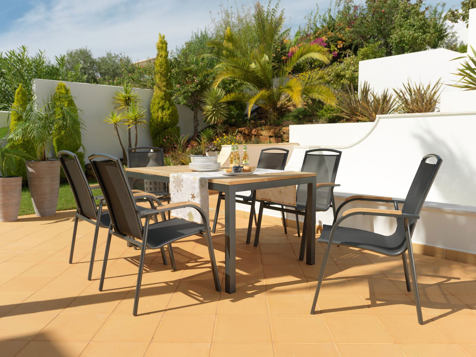 Ambia Garden Ausziehtisch PLANO Moderner Gartentisch mit sechs schwarzen Stühlen auf einer sonnigen Terrasse, umgeben von grünen Pflanzen, aus seitlicher Perspektive.