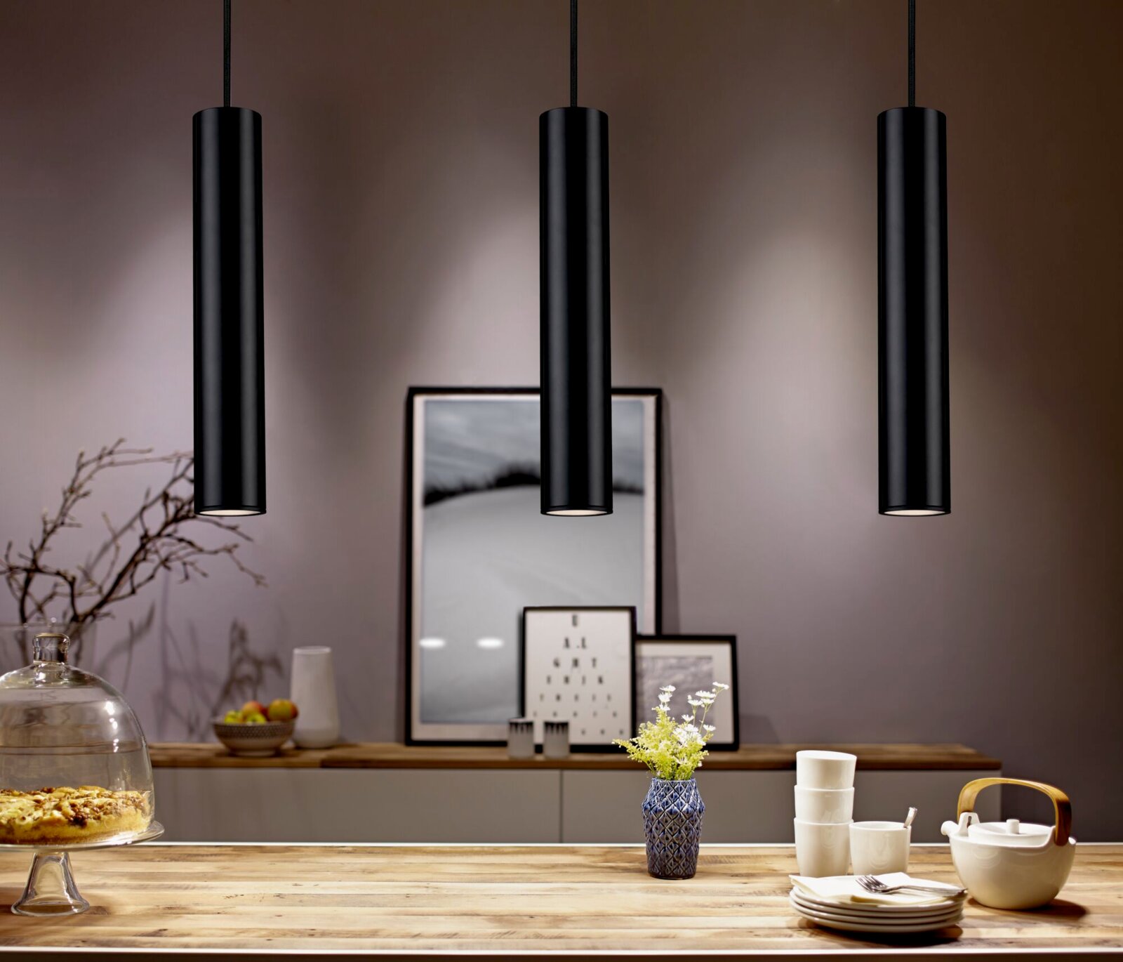 Drie zwarte, cilindervormige hanglampen hangen aan het plafond boven een stijlvol gedecoreerde eettafel. Perspectief: vooraanzicht op de lampen en de tafel.