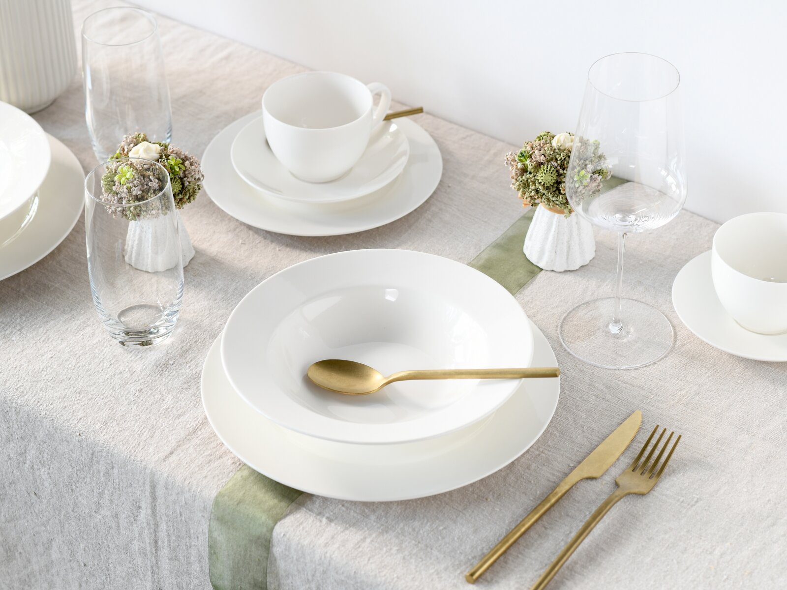 Modern gedeckter Esstisch mit weißem Porzellangeschirr, goldfarbenem Besteck und klaren Gläsern auf Leinentischdecke, schräg von oben fotografiert