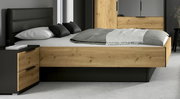 Modernes Doppelbett in Eiche-Optik mit schwarzem gepolstertem Kopfteil und integrierten Schubladen, seitliche Frontansicht im Schlafzimmer-Setting