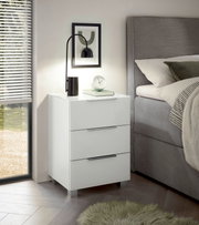 Moderne weiße Nachtkommode mit drei Schubladen, seitliche Perspektive, dekoriert mit einer Lampe und einem Buch, neben einem Bett platziert.