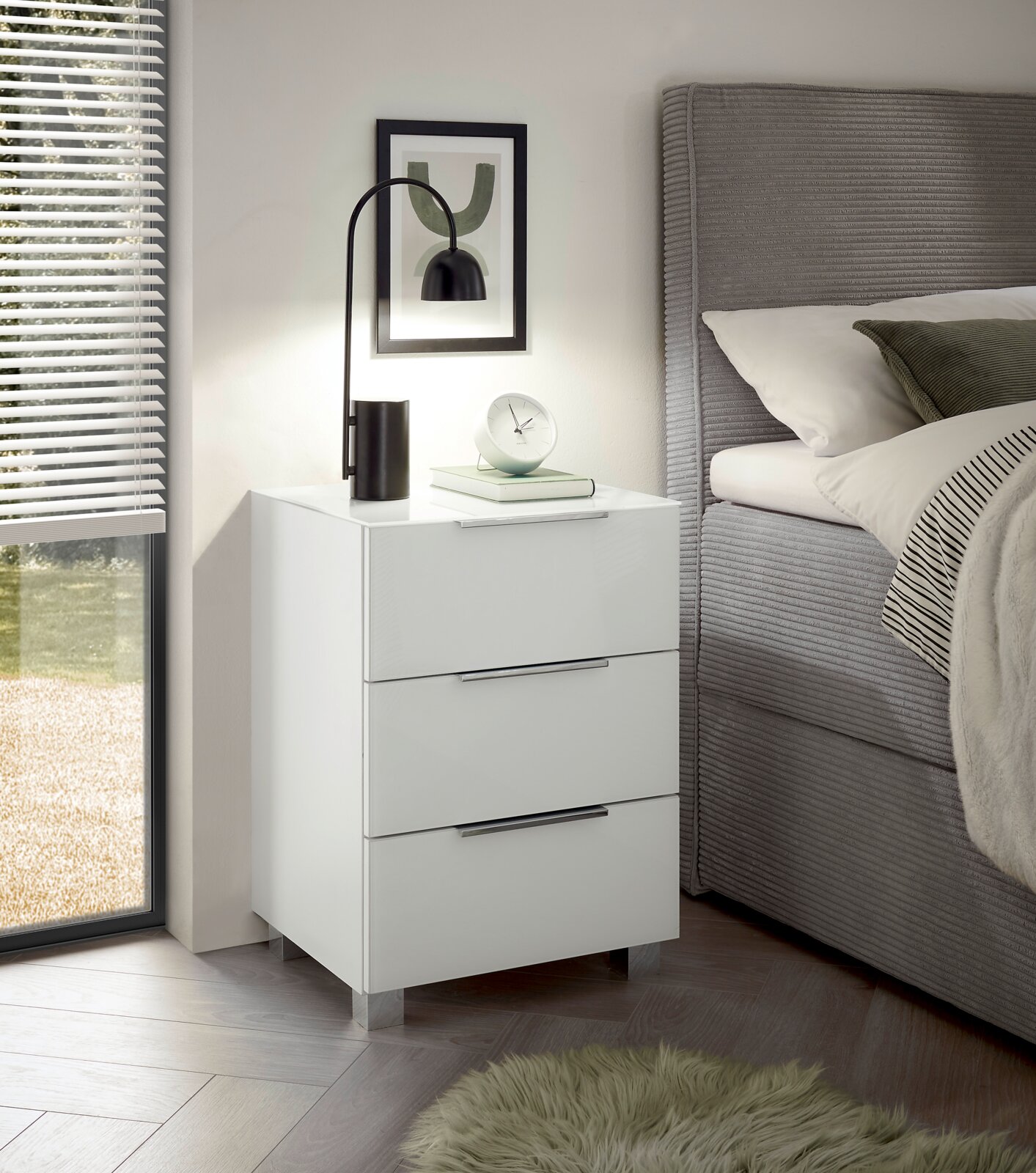 Moderne weiße Nachtkommode mit drei Schubladen, seitliche Perspektive, dekoriert mit einer Lampe und einem Buch, neben einem Bett platziert.