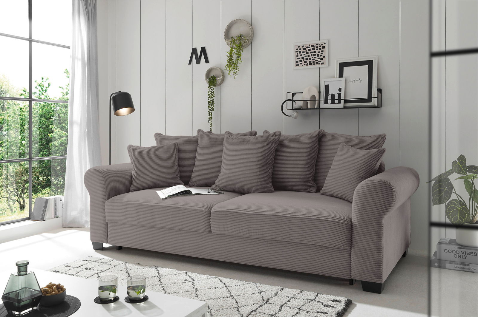 Hom´in Big Sofa MAUREEN Graues Funktionssofa mit mehreren Kissen, seitliche Perspektive, in einem modernen Wohnzimmer mit großen Fenstern und dekorativen Elementen an der Wand.