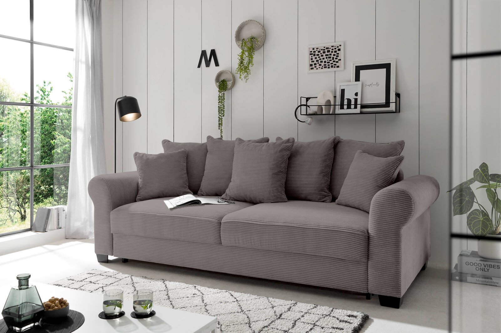 Graues Funktionssofa mit mehreren Kissen, seitliche Perspektive, in einem modernen Wohnzimmer mit großen Fenstern und dekorativen Elementen an der Wand.