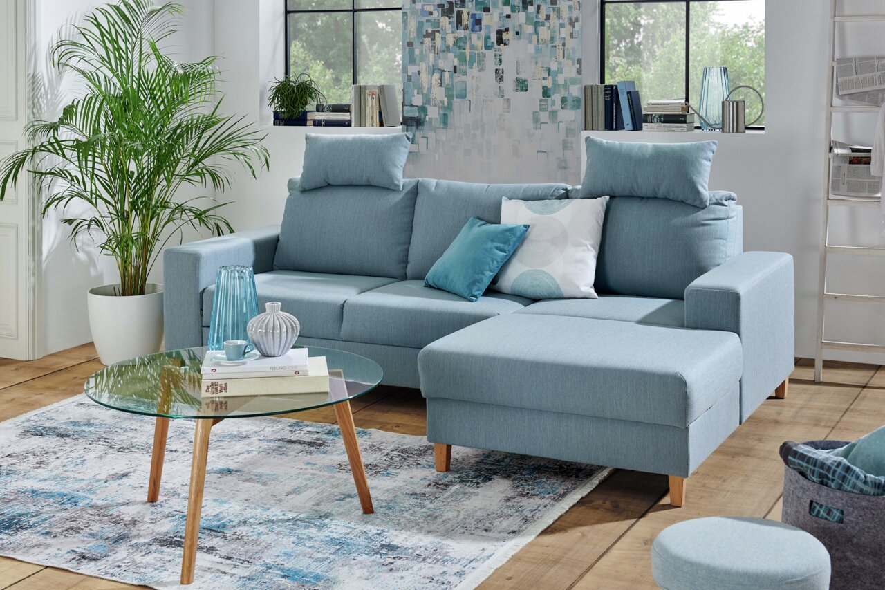 Blaue Eckcouch Ti´me Rundecke ANABELLE in einem modernen Wohnzimmer, mit einem runden Glastisch und dekorativen Kissen, aus der Perspektive von vorne rechts.