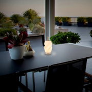 Leuchtende, wellenförmige tragbare LED-Tischlampe in Weiß mit Griff auf einem dunklen Outdoor-Tisch auf der Terrasse, Vorderansicht in leichter Schrägperspektive