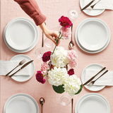 Vogelperspektive auf einen gedeckten Tisch mit weißen Frühstückstellern, Besteck und Blumenarrangement auf rosa Tischdecke.