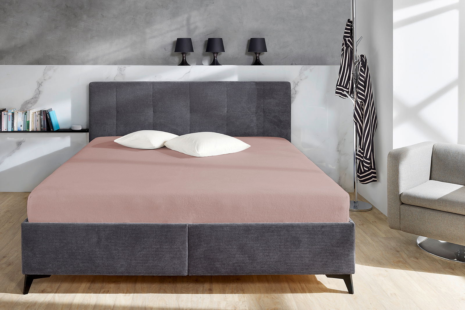 Bett mit grauem Kopfteil und rosa Spannbetttuch, frontal fotografiert, mit zwei weißen Kissen.
