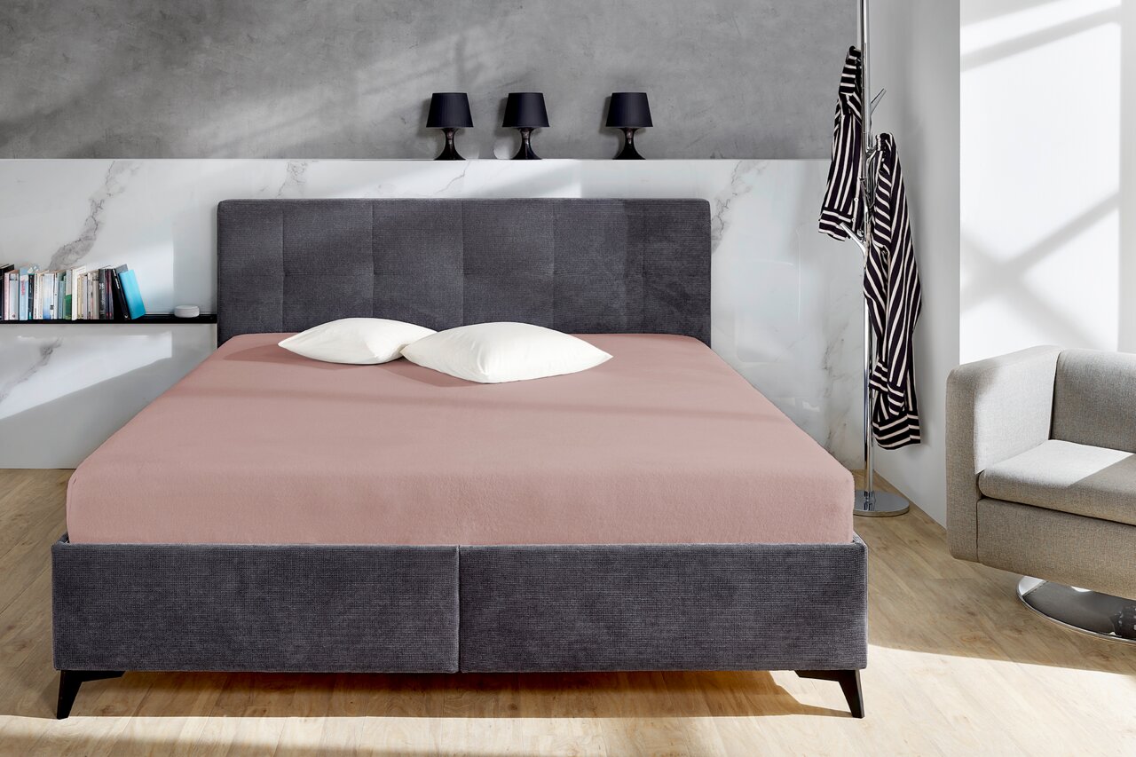 Bett mit grauem Kopfteil und rosa Spannbetttuch, frontal fotografiert, mit zwei weißen Kissen.