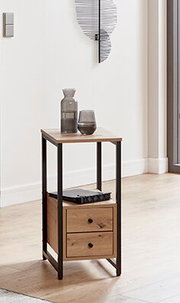 Schmaler Beistelltisch aus Holz mit schwarzem Metallrahmen, zwei Schubladen und Ablage, in heller Wohnraumszene mit Vase und Glas darauf, perspektivisch in leichter Seitenansicht von vorne fotografiert.