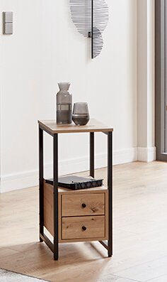 Schmaler Beistelltisch aus Holz mit schwarzem Metallrahmen, zwei Schubladen und Ablage, in heller Wohnraumszene mit Vase und Glas darauf, perspektivisch in leichter Seitenansicht von vorne fotografiert.