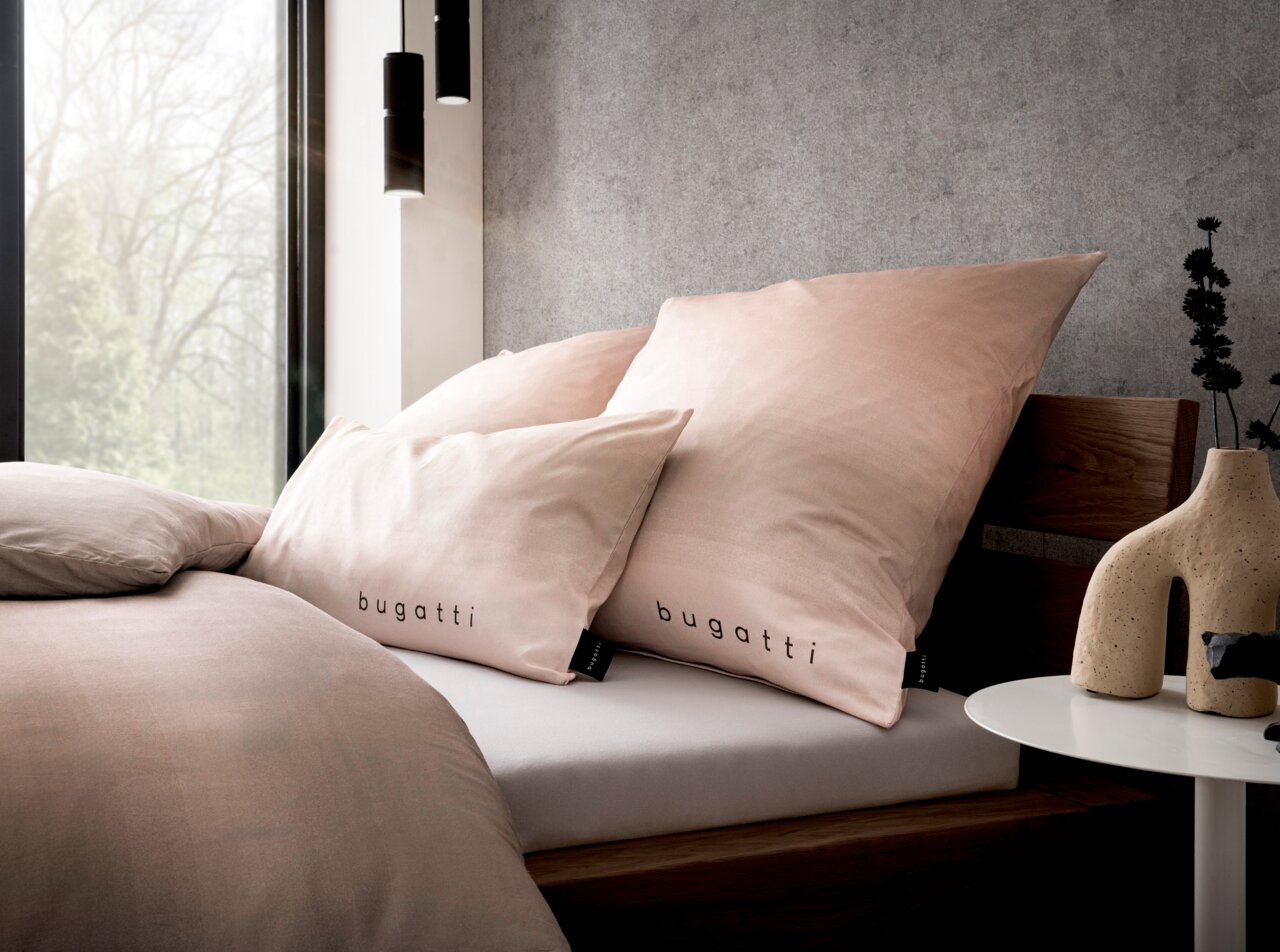 Bettwäsche-Set in sanftem Rosa mit 'bugatti'-Logo, auf einem modernen Holzbett arrangiert, seitliche Perspektive.