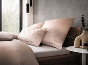 Bettwäsche-Set in sanftem Rosa mit 'bugatti'-Logo, auf einem modernen Holzbett arrangiert, seitliche Perspektive.