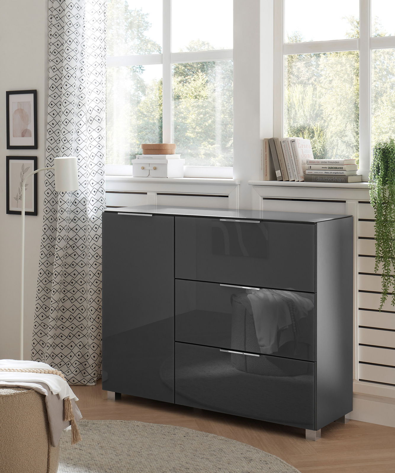 XORA Kommode LIPPO Moderne, dunkelgraue Kommode mit Hochglanzoberfläche, seitliche Perspektive, in einem hellen Raum mit großen Fenstern und dekorativen Elementen.